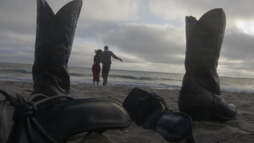 A man and woman playfully run towards the water on a beach as their boots and shoes sit in the sand