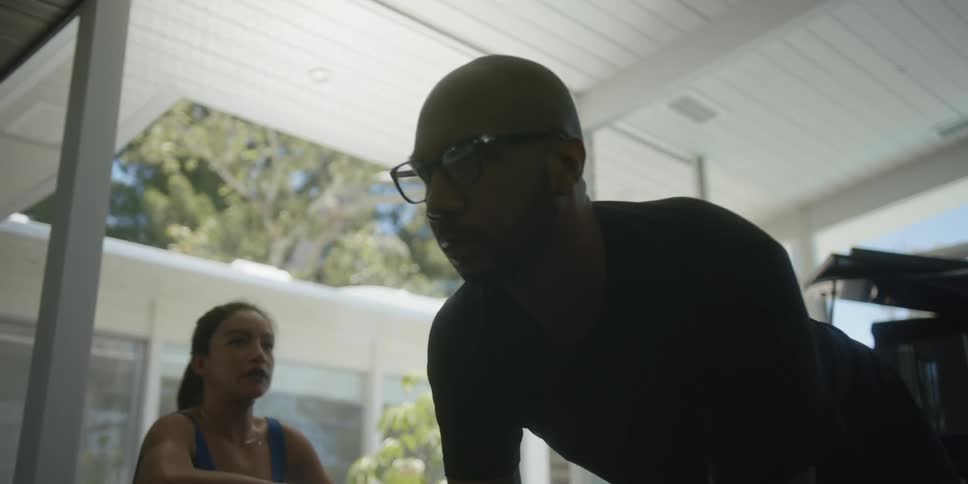 Man with shaved head glasses and black sweatshirt does push ups next to woman with ponytail and blue top in the gym