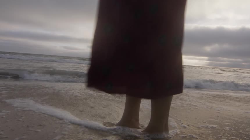 Waves roll over a womans bare feet as she stands in the sand at a beach and the wind blows her sundress under a cloudy sky