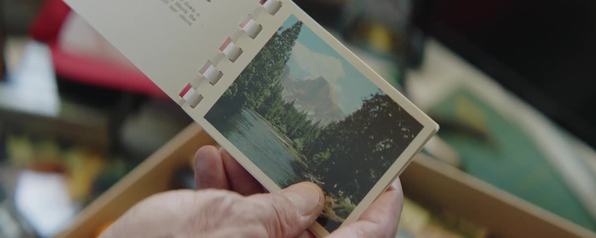 A man is holding a souvenir booklet and looking at a picture of a lake surrounded by forests and a mountain in the distance