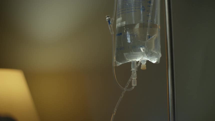 IV drip bag hangs on metal pole near person with lamp in the background