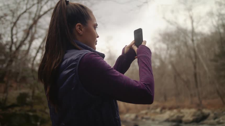 A woman uses her smartphone to film a river running through woodland during the day
