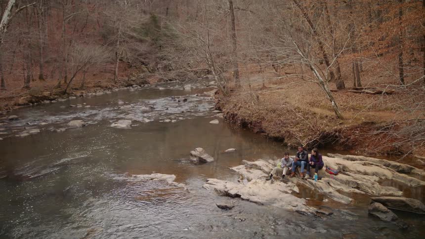 Three people are sitting on rocks along the side of a flowing stream talking and laughing among bare trees and fallen leaves covering the ground