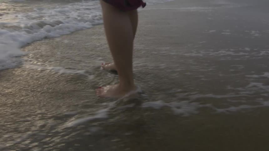 Barefoot woman wearing a dress walks on the shoreline of a beach at evening