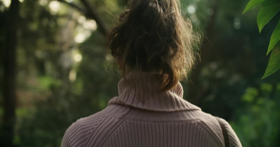 Sunlight shines through trees as a woman wearing a sweater is walking through neighborhood woods