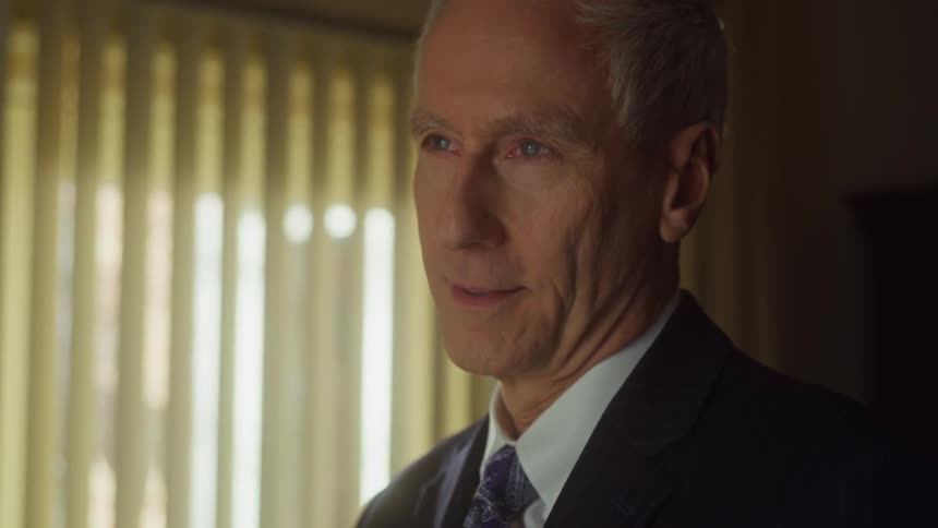 A man wearing a suit is looking down with a smile on his face and he looks up as the sun shines through the curtains