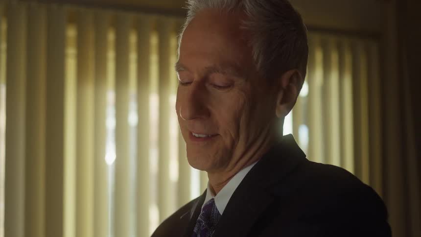 A man is looking down with a smile on his face and he looks up as the sun shines through the curtains