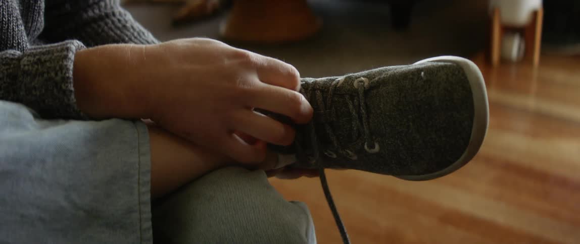 A girl is sitting down and she puts her foot on her knee and unties her shoe and takes it off