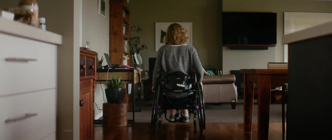 A woman in a wheelchair propers herself through a house to reach a laptop on a nearby desk