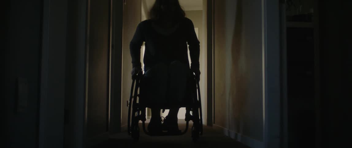 A girl in a wheelchair rolls down a hallway and into a room as she holds the wheels with her hands and moves forward