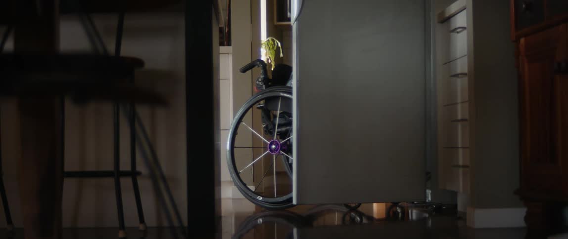 A woman at home wheels her wheelchair to the fridge and gets out some orange juice and closes the door and wheels away