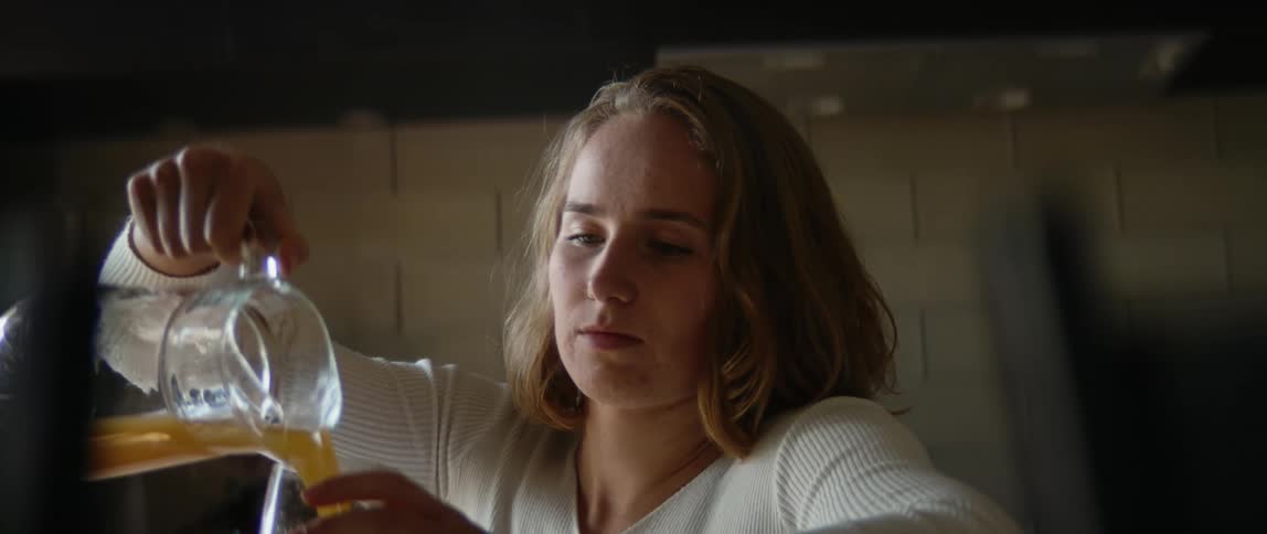 Blonde female wheelchair user wearing white shirt pours juice into glass in the kitchen