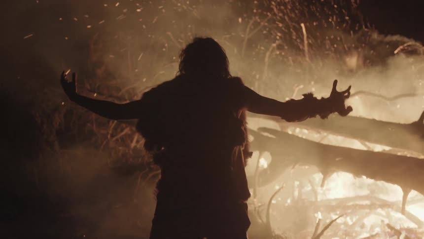 A man is standing by a fire and he is raising his arms