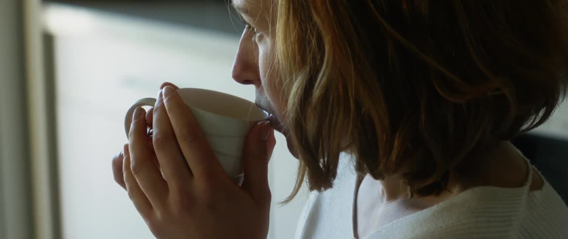 Redheaded woman drinks from cup in kitchen while looking out window