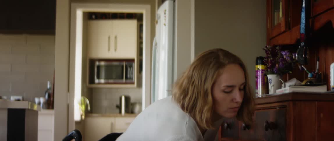 A girl in a wheelchair rolls into the kitchen with a dog food bowl in her lap and she bends over and sets it on the floor