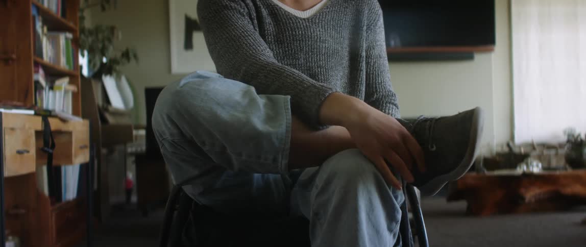 Blonde female wheelchair user wearing gray sweater white socks and jeans wears shoes in the living room