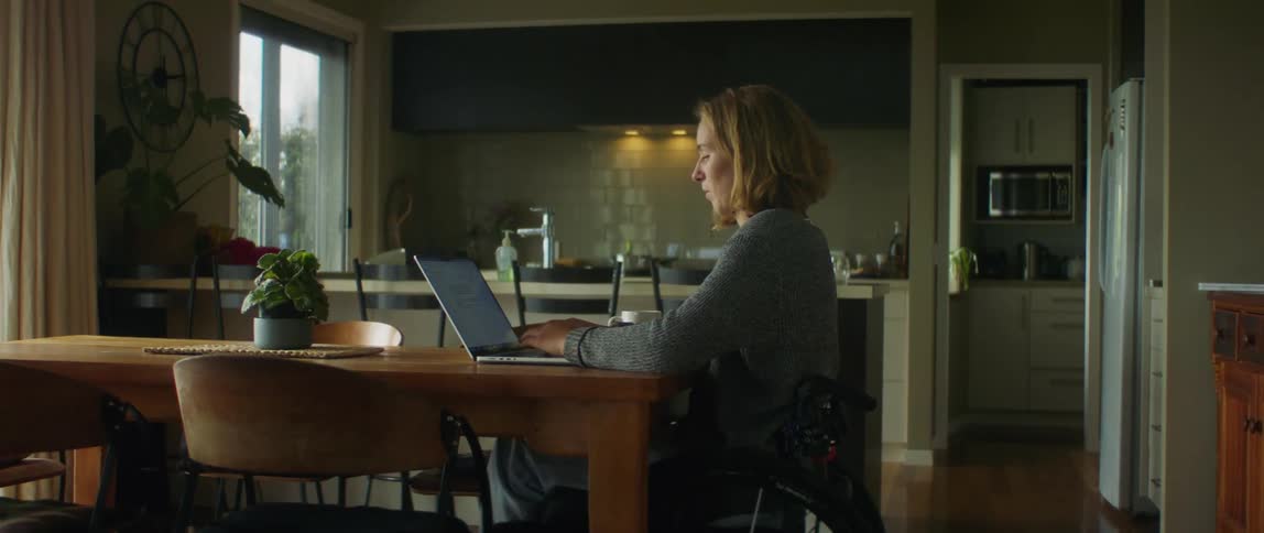 Woman sits in wheelchair at dining table in home and types on laptop