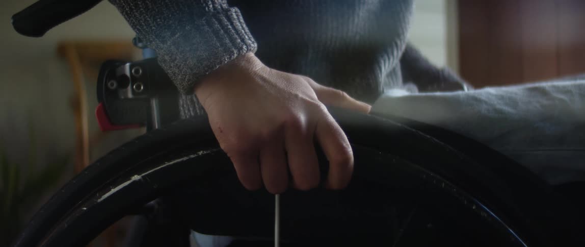 Woman sitting on a wheelchair grips one of the wheels with one hand in the living room