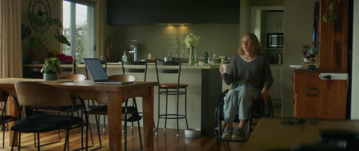Woman rolls wheelchair to work at laptop after filling mug with water at kitchen sink