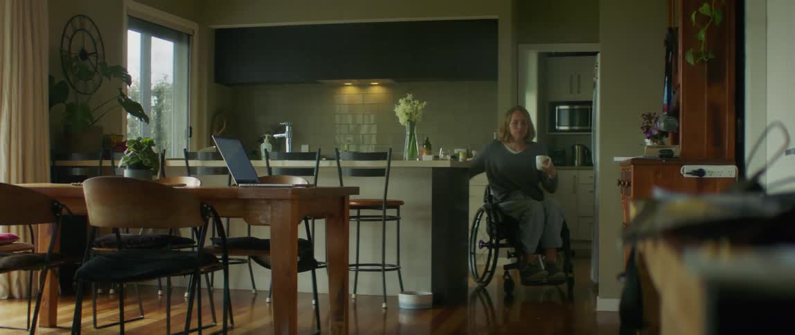Woman in wheelchair fills cup from kitchen faucet before returning to laptop on dining table