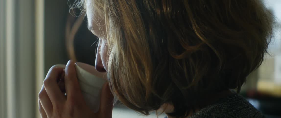 A woman drinks from a steaming coffee mug while sitting indoors during the day