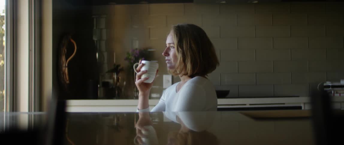 Woman sits in kitchen drinking from a mug and looking out the window