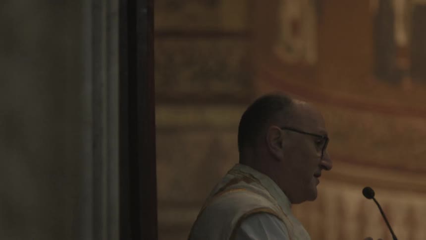 A priest stands at a microphone in a church with saints painted on the walls
