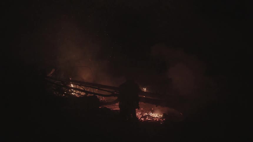 A person stands before a large fire at night