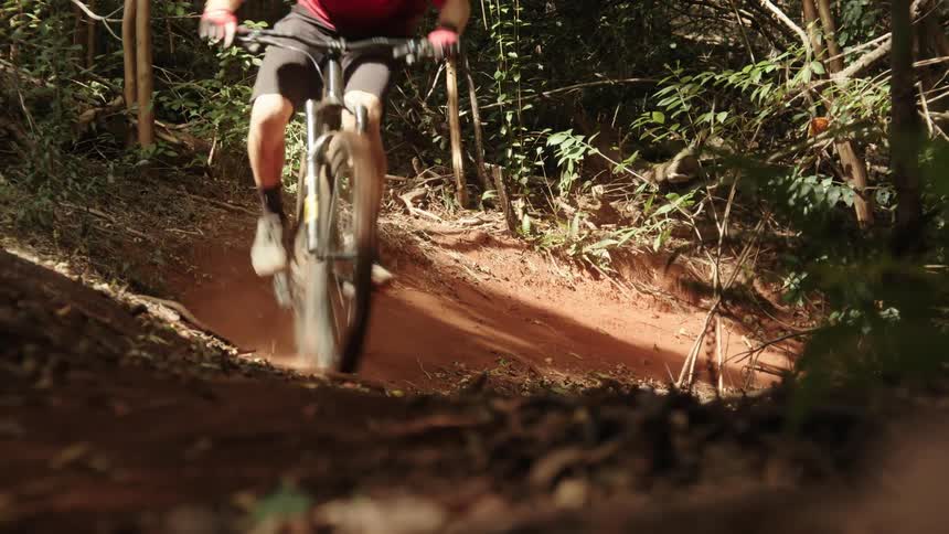Cyclists rides bike over red dirt trail through woods and kicks up dust