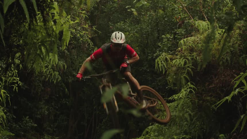 A boy on a bicycle rides up a dirt hill on a trail in the woods and leaps into the air and turns the bike before he lands