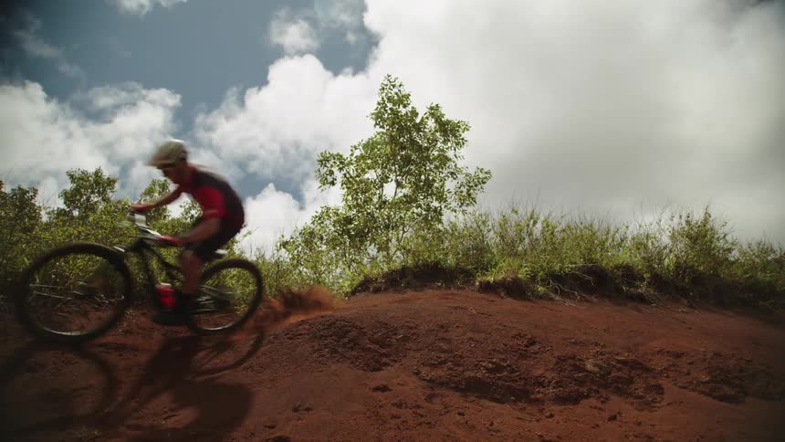 A boy on a bicycle rides up a dirt hill near trees and leaps into the air under a cloudy sky