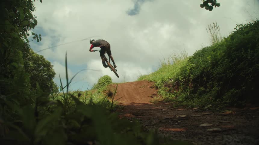 A boy on a bicycle rides up a dirt trail on a hill and leaps into the air under a cloudy sky