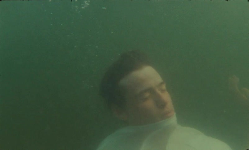 A young man purposely falls underwater in the ocean and stays under with his eyes closed just floating in one spot