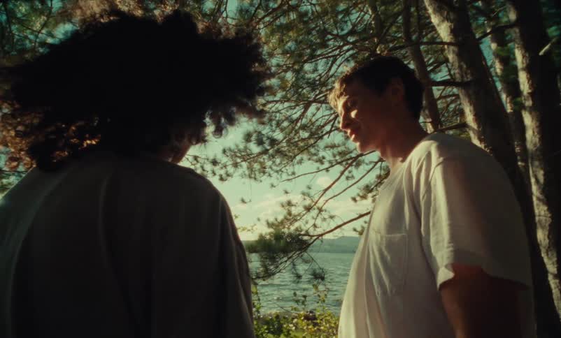 A teenage male and female stand in the woods near a lake and have a conversation