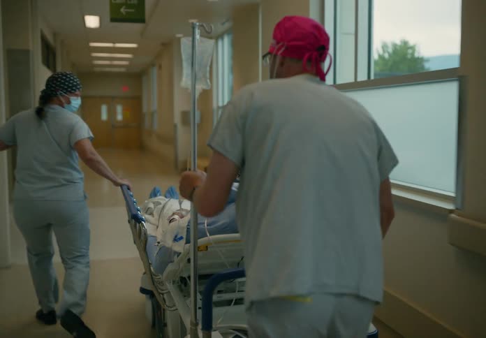 A male and female nurse quickly wheel an old man on a gurney down a hospital hallway and into a room