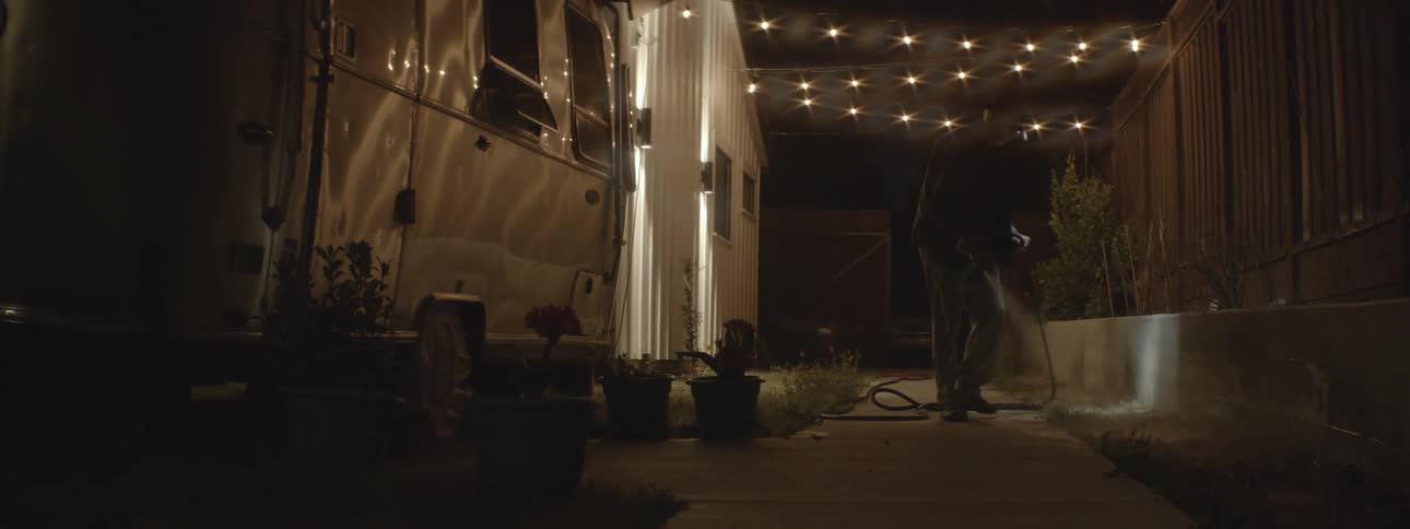 A man wearing a headlamp and carrying a garden hose is walking on a sidewalk watering plants at night