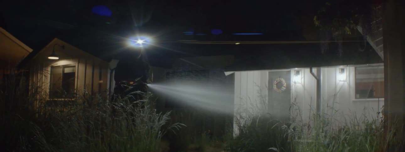 A man is standing outside of his house at night with a garden hose watering plants