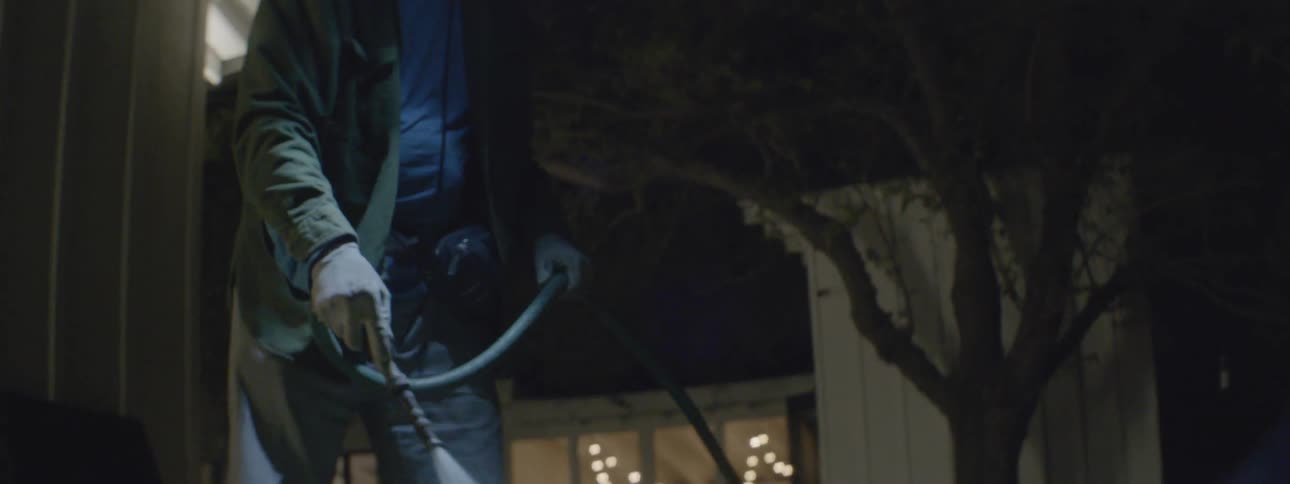 A man is standing outside of his house at night with a garden hose watering plants