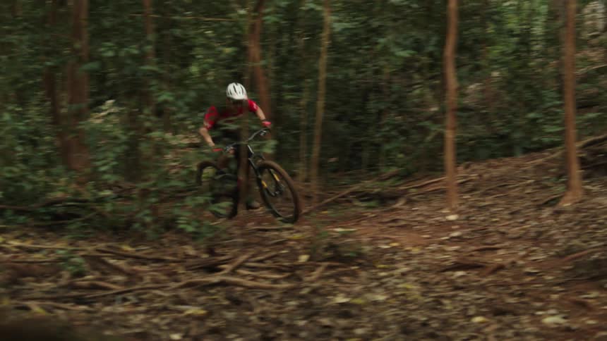 A cyclist rides along a narrow dirt trail through a woodland at high speed during the day