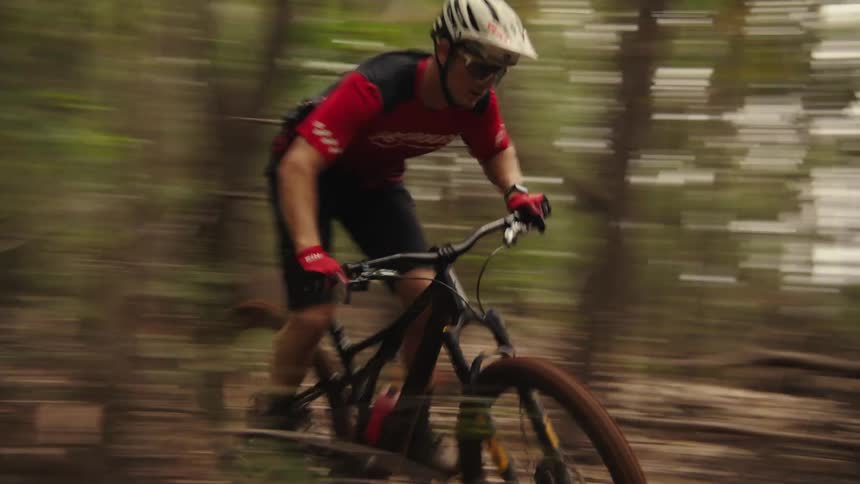 A boy rides his bicycle on a trail in the woods and flies into the air when he goes up a hill