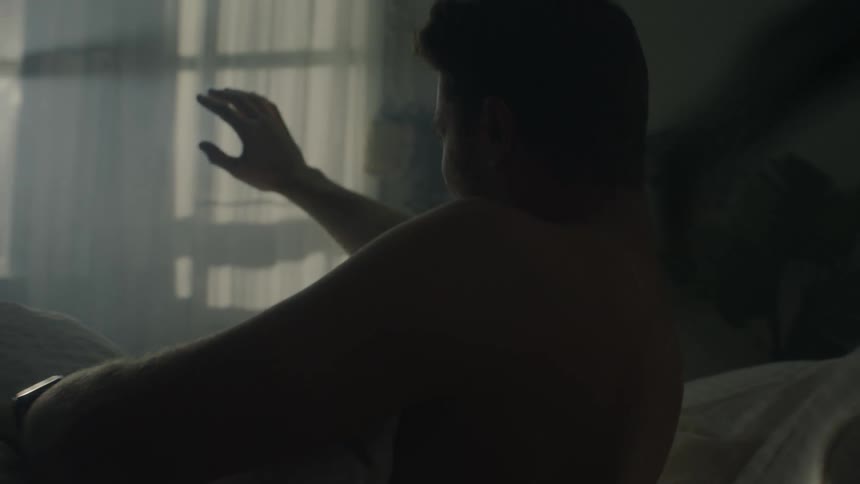 A man is lying in bed and he sits up as light shines through the curtains