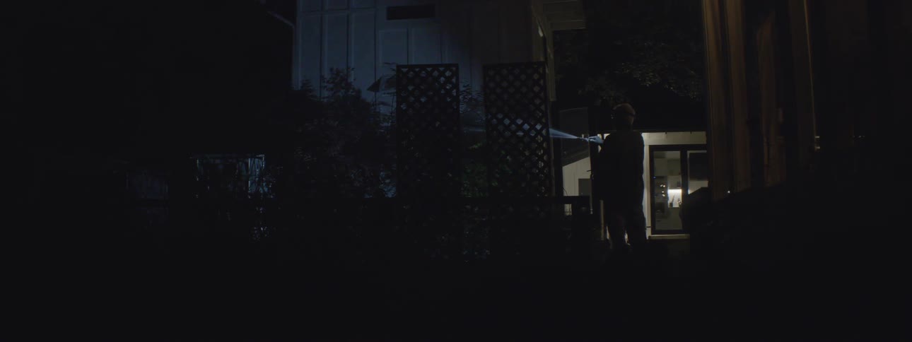 Older man with headlamp waters plants at night before noticing another light shining across his yard