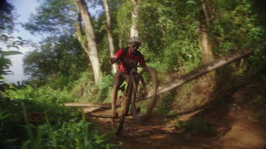 A man rides his bike quickly down a dirt trail and jumps over a large tree trunk blocking the path