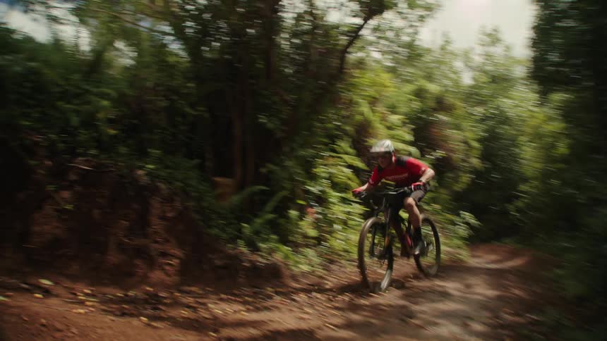 A male mountain biking on a dirt trail in a forest does a jump over a hill