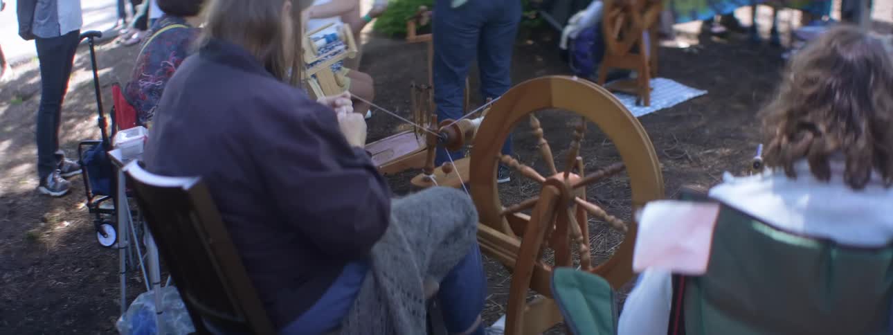 A group of people outside while several women sit and spin wool