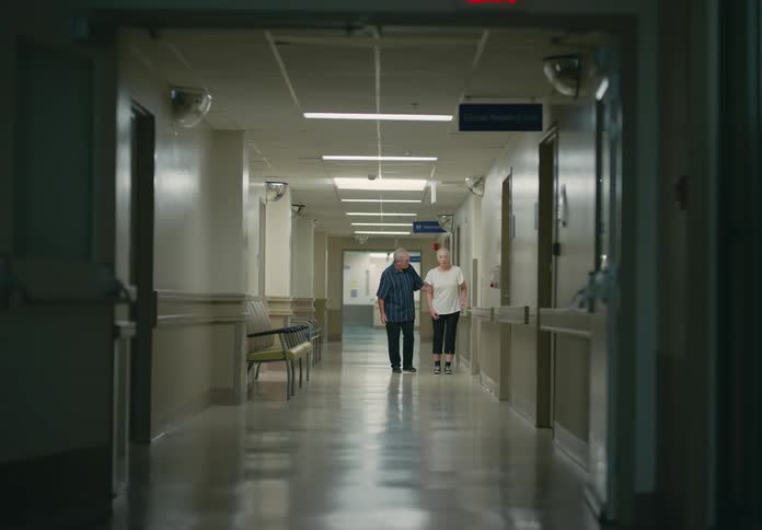 An elderly man helping his elderly wife walk down a hospital hallway