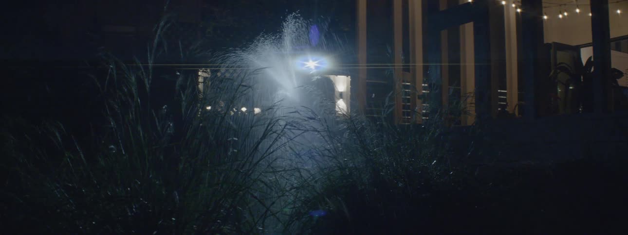 A man wearing a headlamp is standing outside of his house at night with a garden hose watering plants
