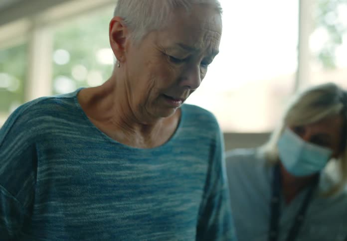 An elderly woman attempts to walk using walking rails in a hospital while a nurse guides her from the side