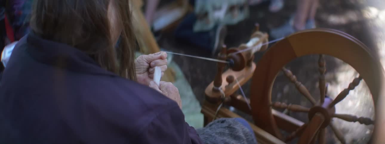 A woman feeds a thread into a spinning wheel while sitting outdoors during the day
