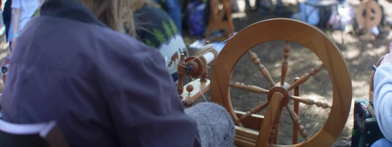 A woman operates a spinning wheel while sitting outdoors during the day